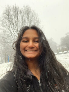 Headshot of a woman with brown skin tone, long black hair, and a black coat against a wintry, snowy background.